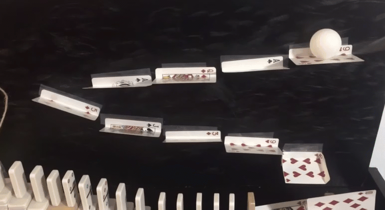 a ball rolls down a ramp made of playing cards and causes a line of dominos to fall