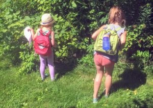 Two young girls explore bushes while wearing backpacks.