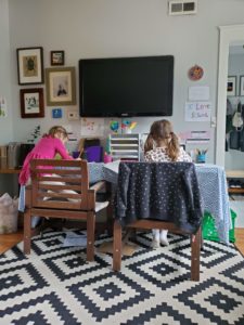 Two young girls do school work at a table set up at home for remote schooling.