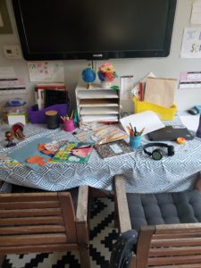 A messy remote schooling desk, full of crafts, books, and notebooks.