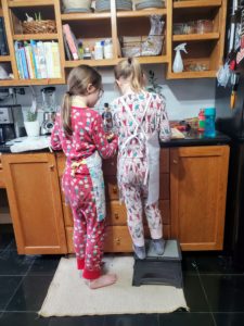 Two young sisters wear aprons over their pajamas to make food in the kitchen.