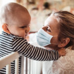 A woman wears a mask while caring for a child in a crib.