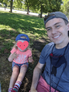 A dad poses for a picture with his daughter while sitting outside at a park.