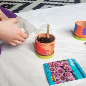 A child uses a spray bottle to water a plant in a painted pot.