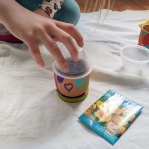 A child places a clear plastic cup over a pot with dirt and seeds in it.